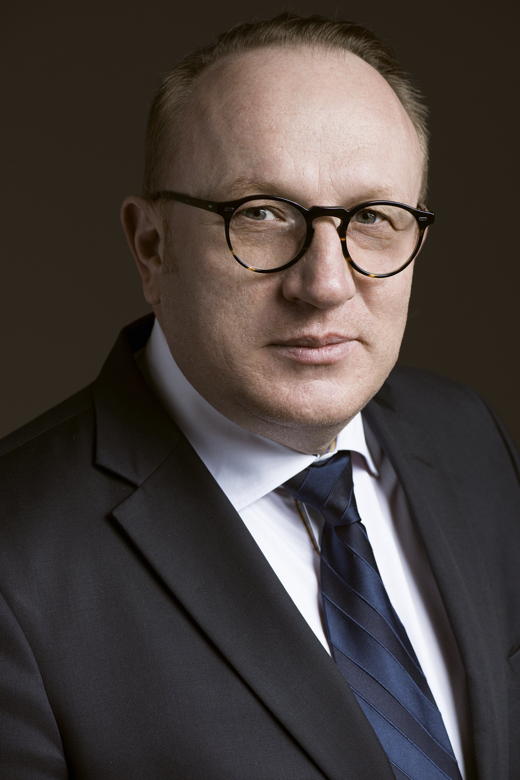 Profesjonalny portret biznesowy mężczyzny w okularach, w czarnym garniturze, białej koszuli i ciemnoniebieskim krawacie, na neutralnym ciemnym tle.